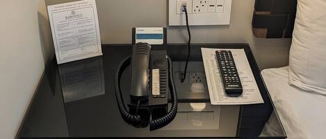 A hotel room with a black bedside table, a lamp, a telephone, a remote control, and a bed with white bedding.