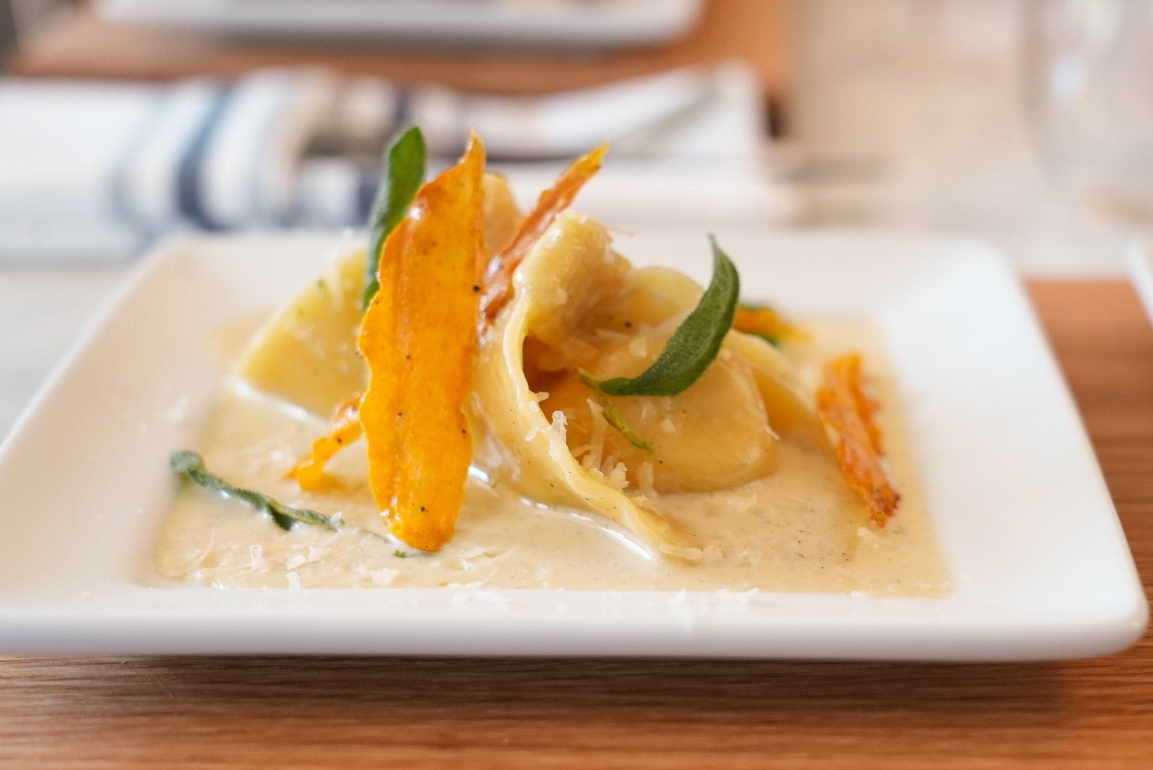 Ein quadratischer weißer Teller mit cremiger Soße, Pasta und Garnituren auf einem Holztisch.