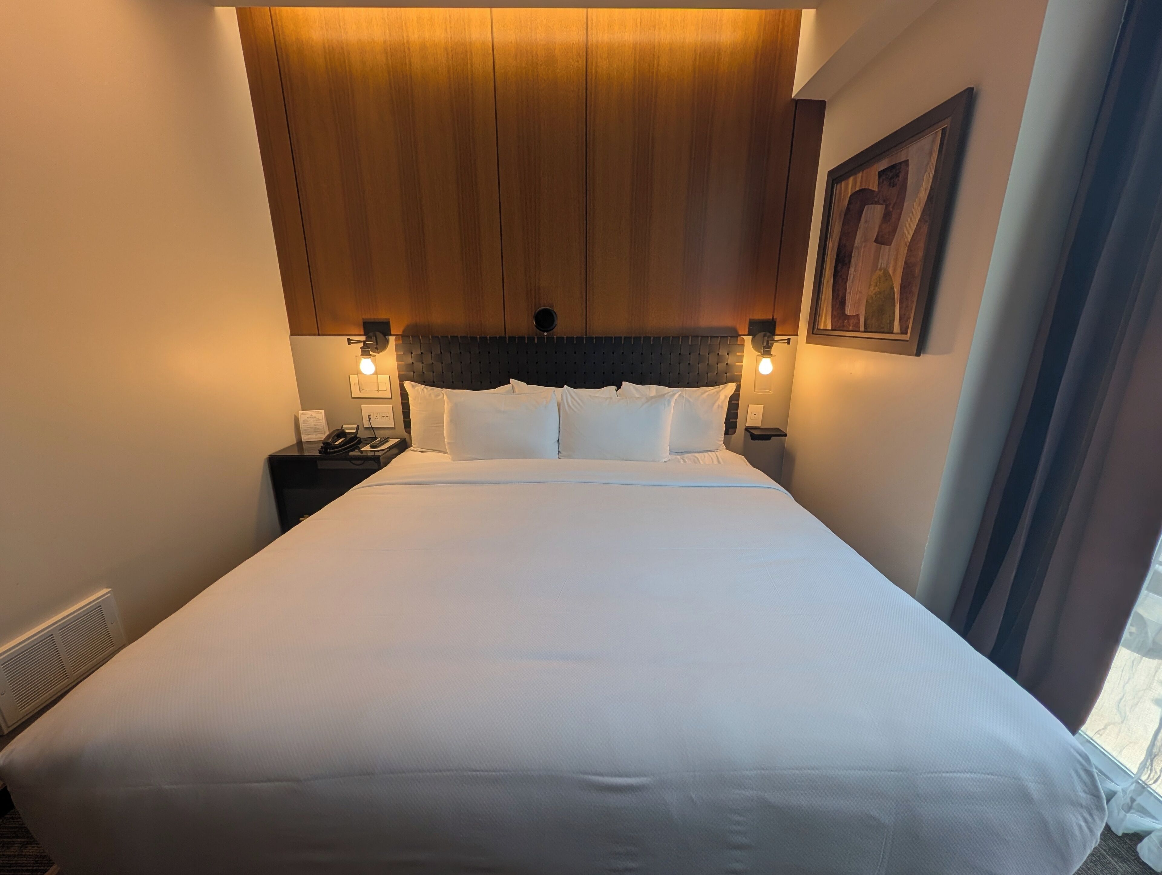 A hotel room with a large bed, wooden headboard, two bedside lamps, a nightstand, and a framed picture on the wall.