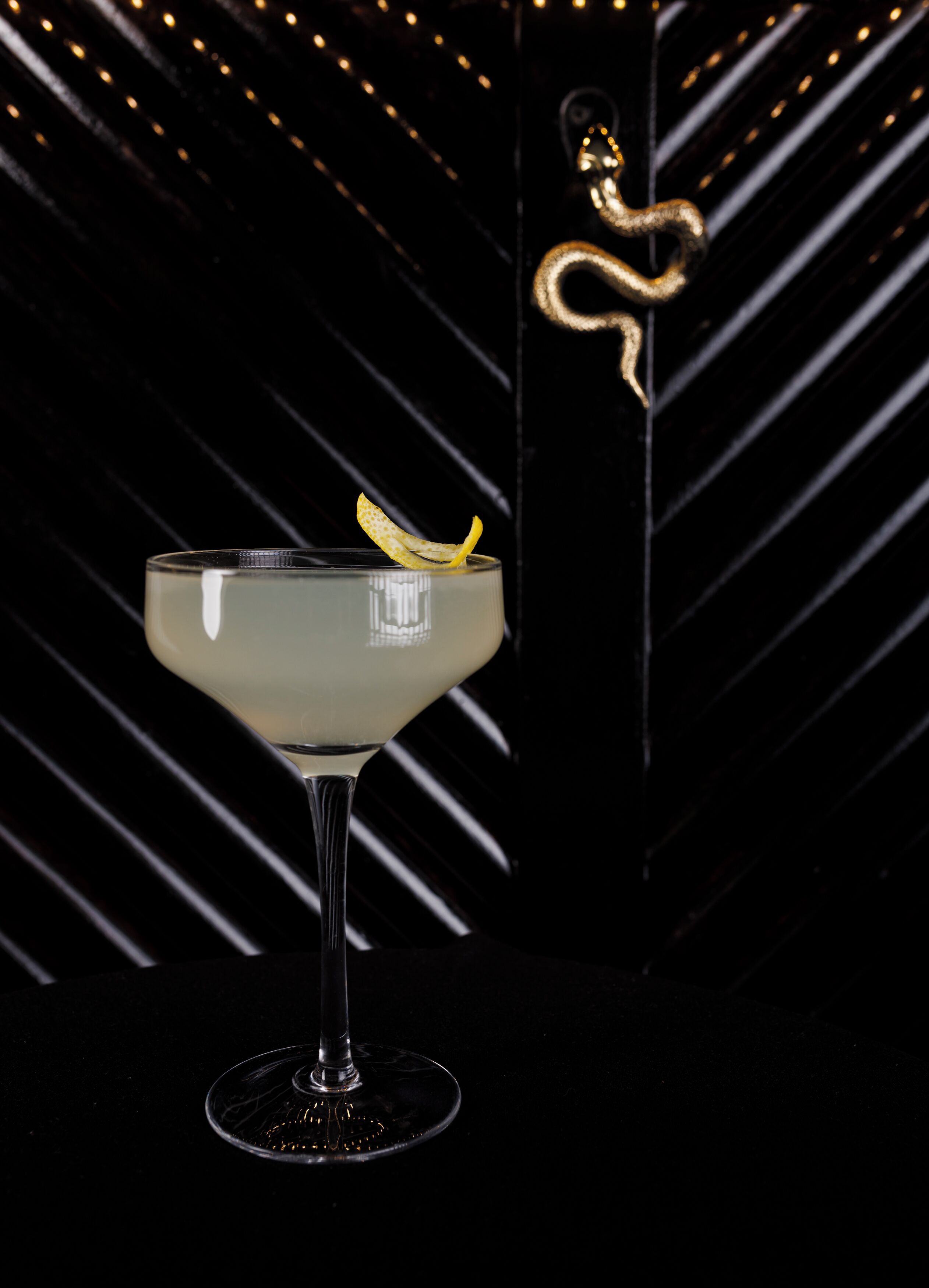 A cocktail in a martini glass with a lemon peel garnish, set against a dark background with diagonal lines and a snake decoration.