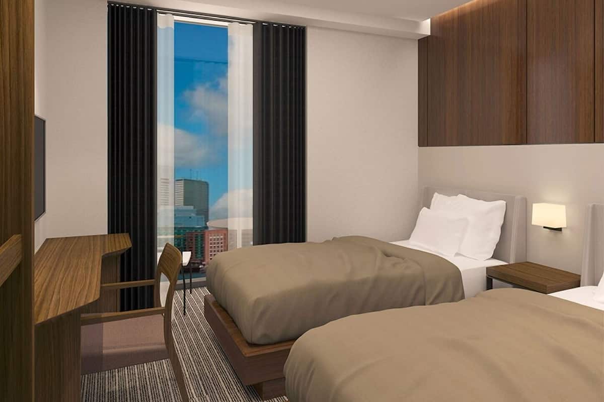 Habitación de hotel con dos camas, un escritorio, una silla, ventana con cortinas y vista a la ciudad.