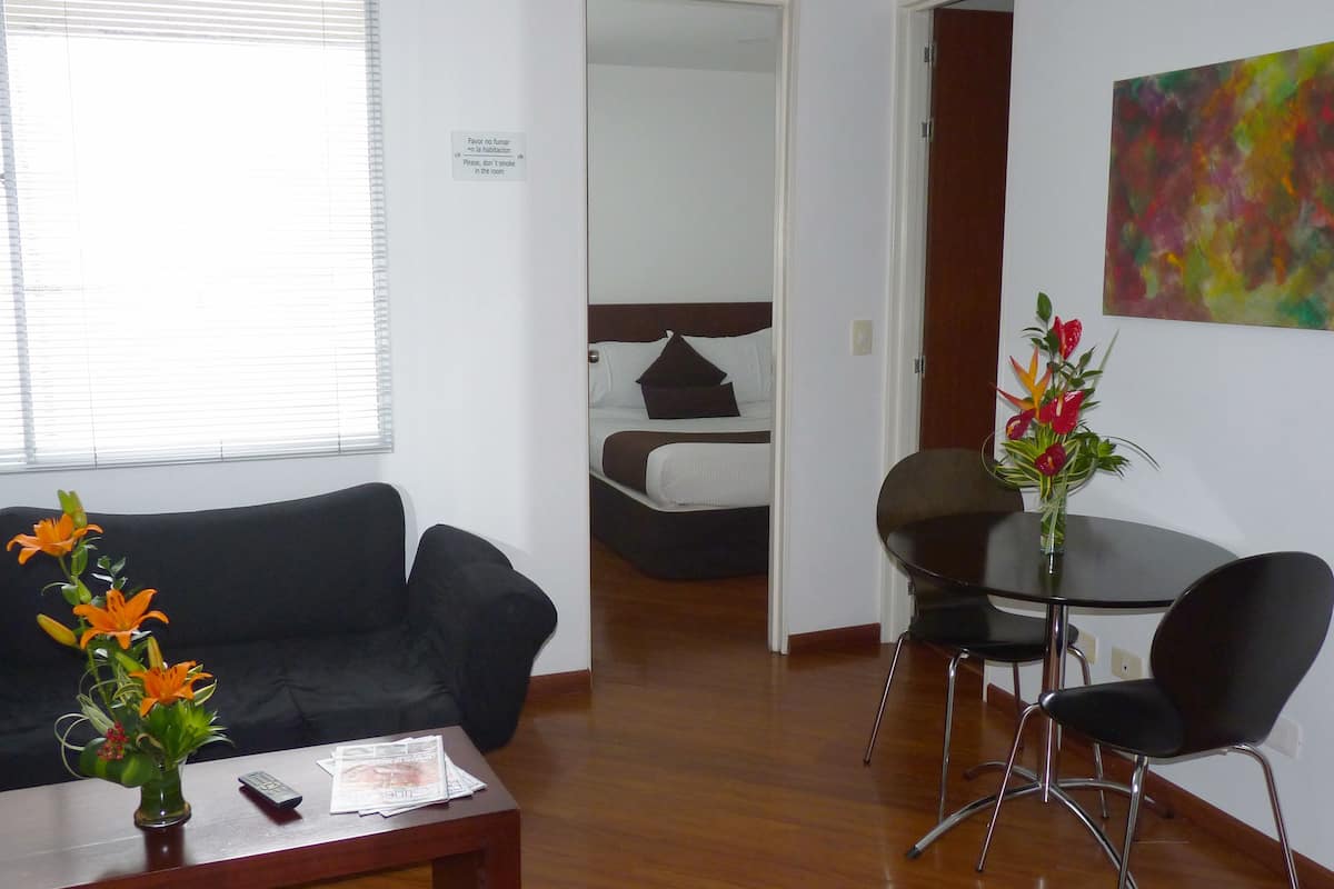Une chambre d’hôtel avec un canapé noir, une petite table sur laquelle est posé un vase de fleurs, une table à manger avec deux chaises, un lit avec une literie blanche et une peinture abstraite colorée accrochée au mur.