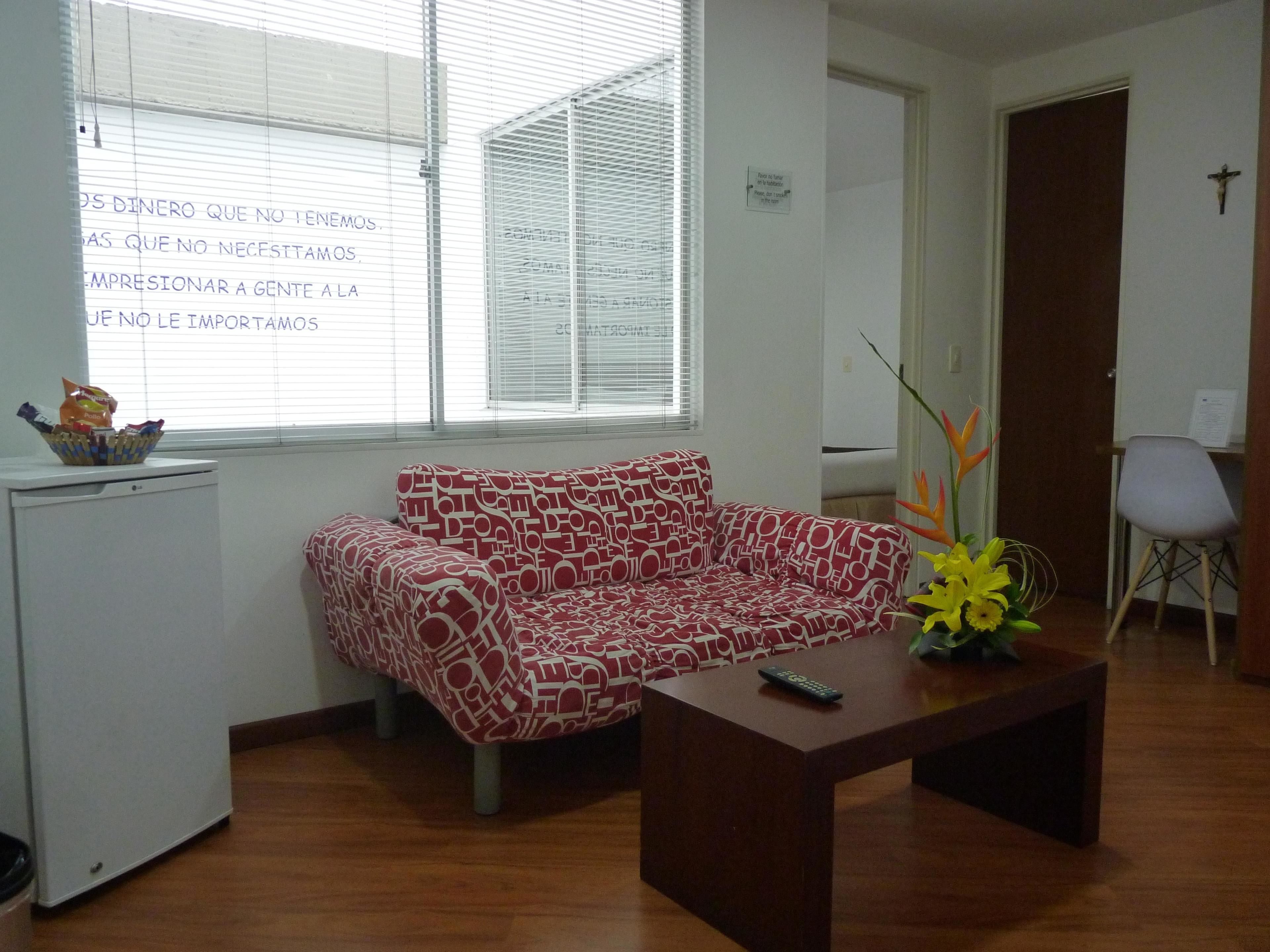 Una habitación pequeña y amoblada con un sofá, una mesita de centro, un refrigerador compacto y una ventana con persianas.
