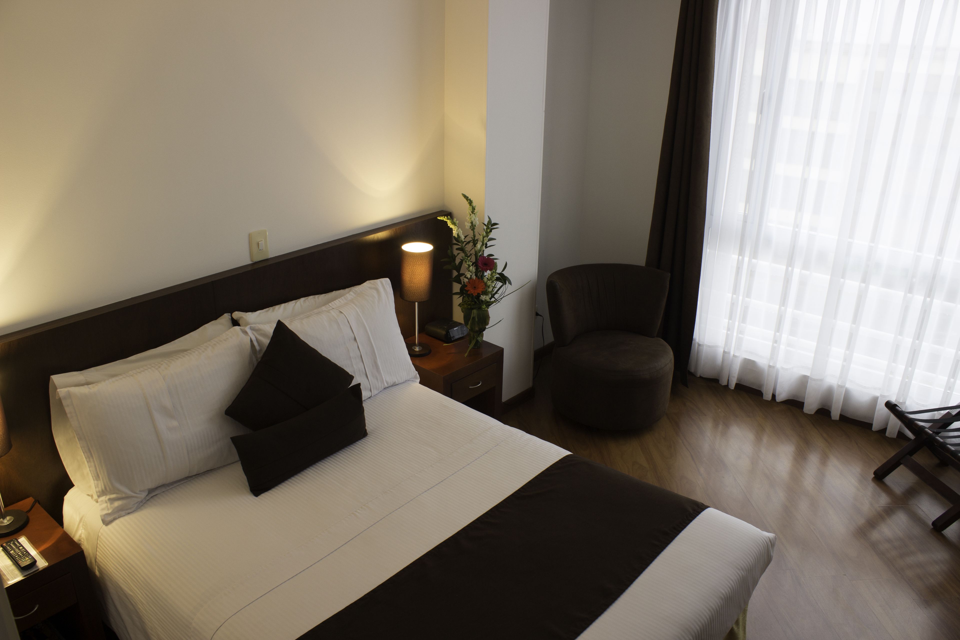 Habitación de hotel con cama, mesita de noche, lámpara, silla y ventana con cortinas.