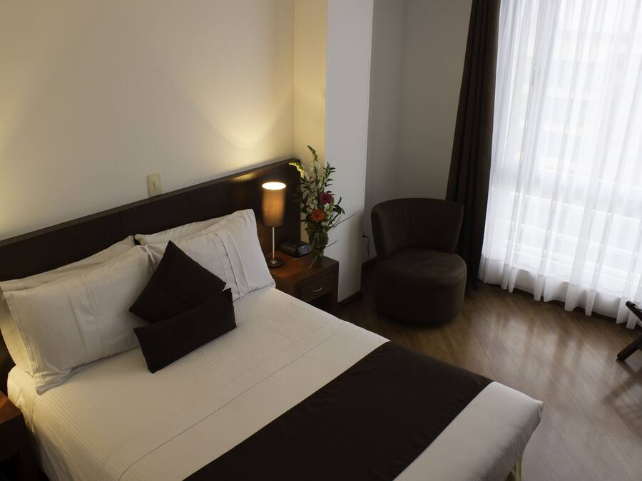 Habitación de hotel con cama, mesita de noche, lámpara, silla y ventana con cortinas.