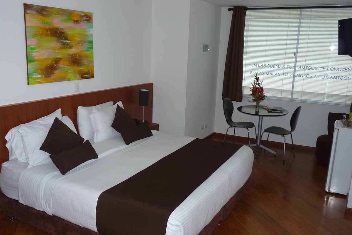 Une chambre d’hôtel avec un grand lit, un bureau, deux chaises, une petite table et une fenêtre avec des stores.