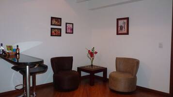 A room with a bar area, two chairs, a small table with a vase of flowers, and three framed pictures on the wall.
