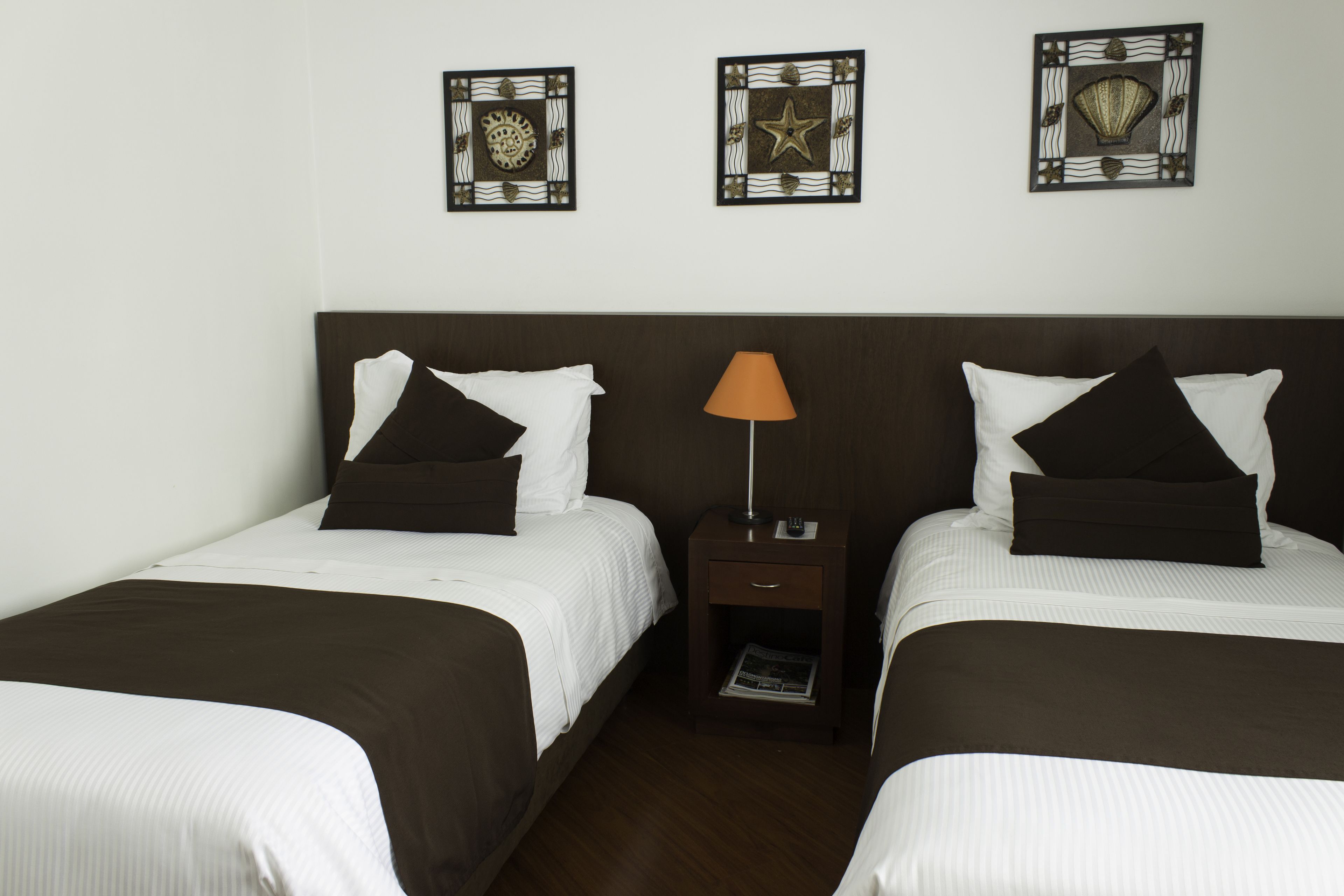 Habitación de hotel con dos camas, cada una con ropa de cama blanca y cabeceros marrón oscuro. Hay obras de arte decorativas sobre las camas y una mesita de noche pequeña con una lámpara entre ellas.