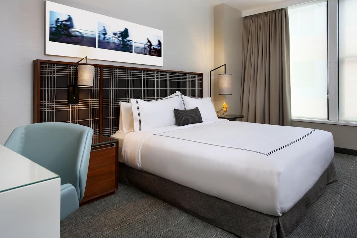 A hotel room with a large bed, a desk, a chair, a headboard with a grid pattern, and a window with curtains.