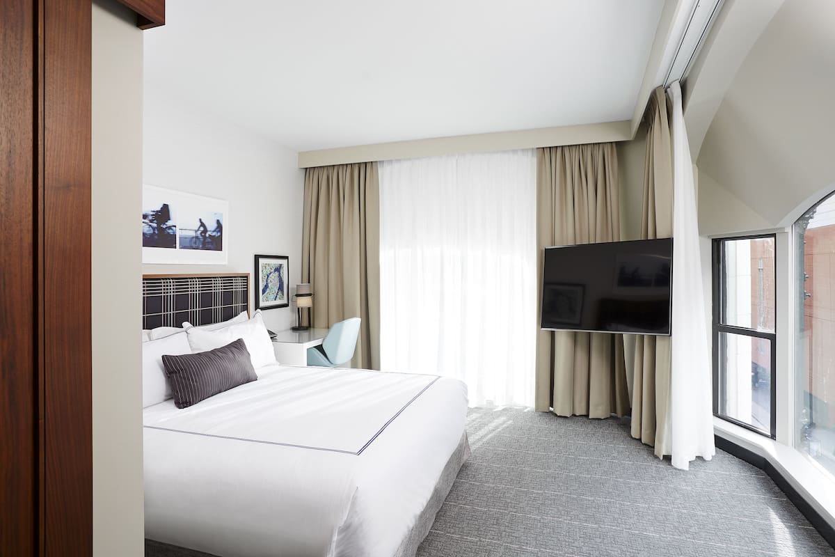 A hotel room with a large bed, a desk with a chair, a flat-screen TV, and a large window with curtains.