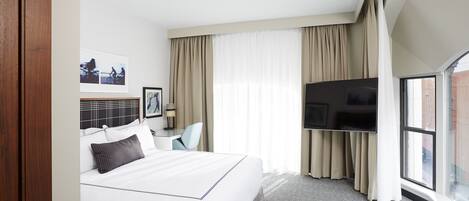 A hotel room with a large bed, a desk with a chair, a flat-screen TV, and a large window with curtains.