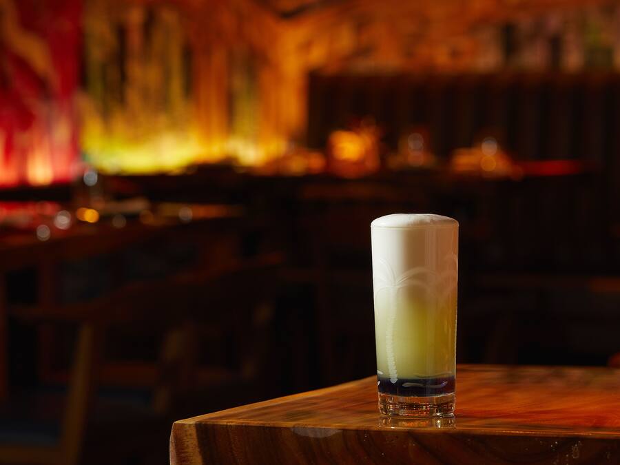 A tall glass of a creamy beverage on a wooden table in a dimly lit bar with wooden furniture and a blurred background.