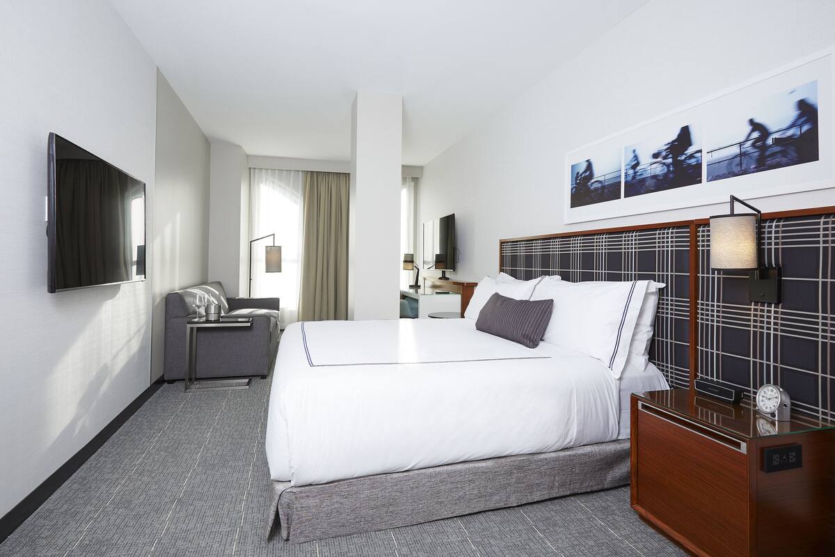 A hotel room with a large bed, a TV mounted on the wall, a desk with a lamp, and a clock on the nightstand.