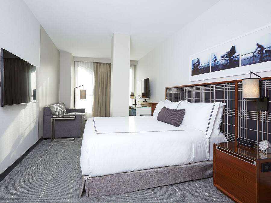 A hotel room with a large bed, a TV mounted on the wall, a desk with a lamp, and a clock on the nightstand.