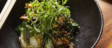A black bowl with a dish of food, including greens and possibly meat, garnished with sesame seeds.