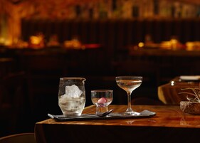 A bar setting with a glass of ice water, a small glass with a pink garnish, and a martini glass on a wooden table.