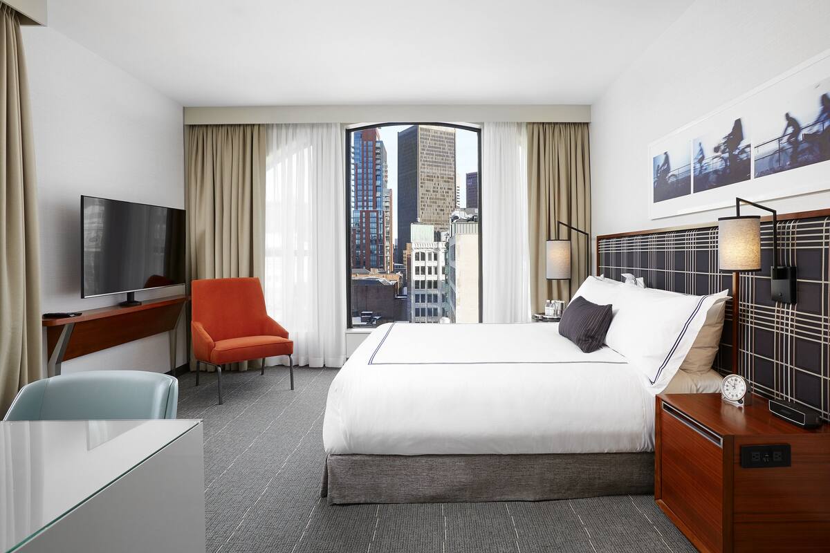 A hotel room with a large bed, a desk, a TV, a chair, and a view of the cityscape.