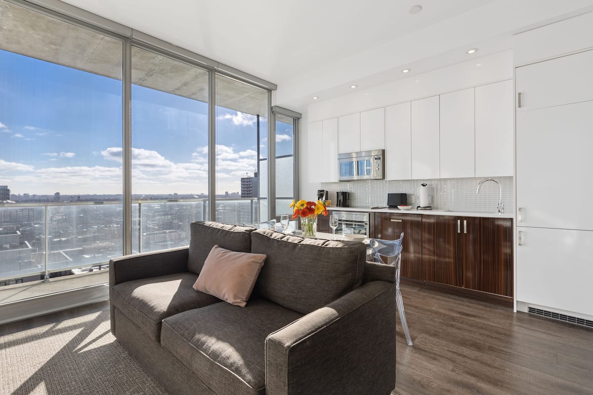 A modern living room with a large window, a sofa, and a kitchen area with white cabinets and a microwave.