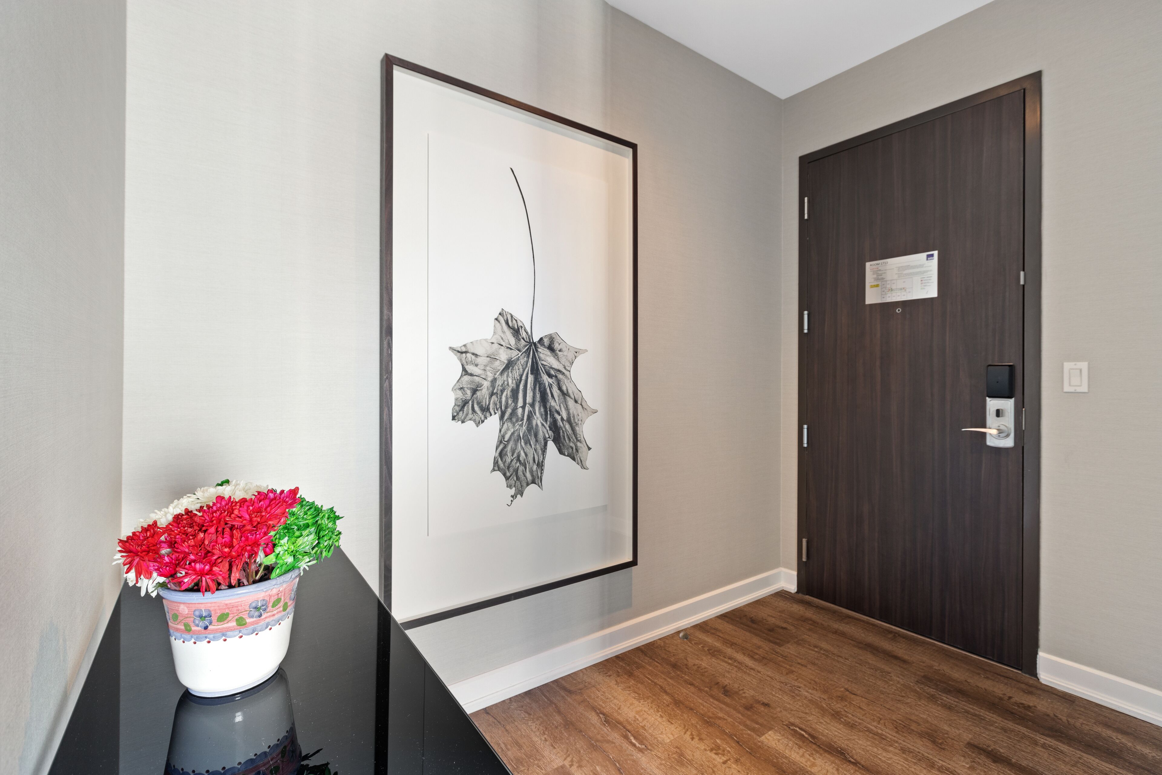 A hallway with a framed botanical print on the wall, a potted plant with red and white flowers, and a dark wooden door with a lock.