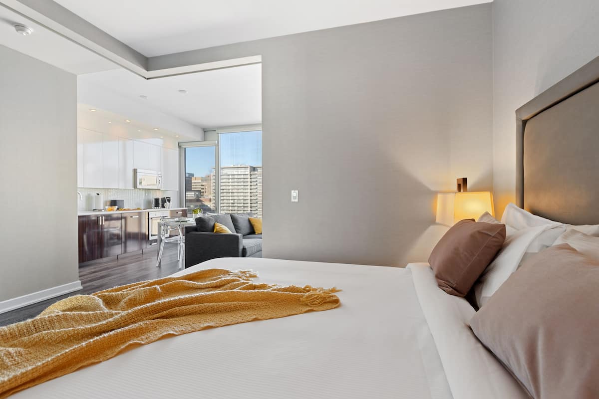 A modern hotel room with a large bed, a view of the city, a kitchenette, and a dining area.