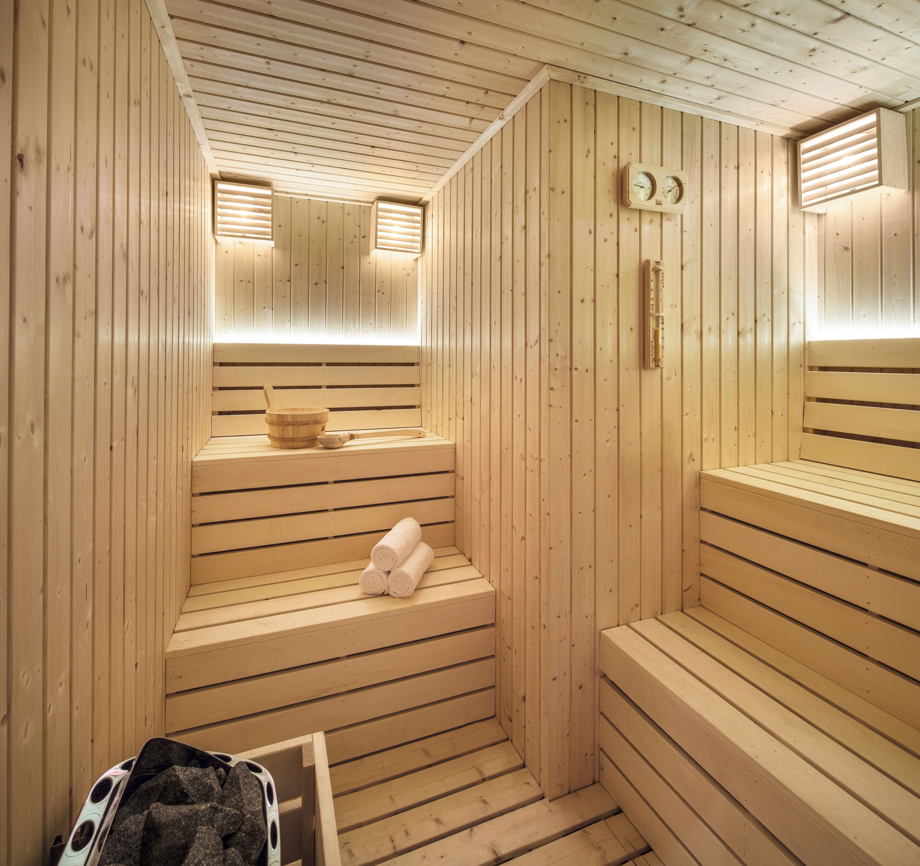 A sauna with wooden benches, a bench with towels, a wooden bucket, and a wall-mounted thermometer.