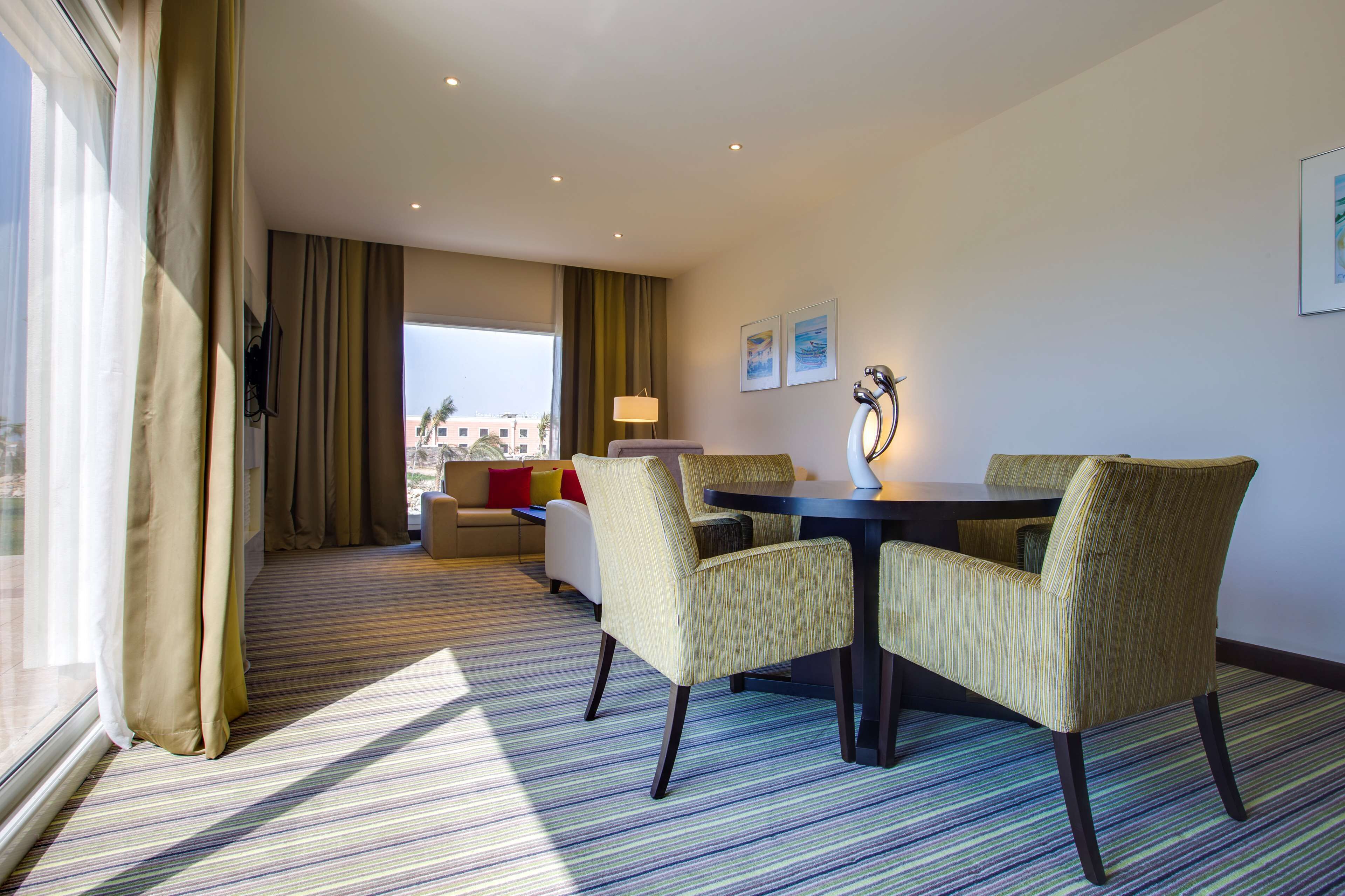 A hotel room with a dining area featuring a round table and four chairs, a sofa, a lamp, and a view of the cityscape through large windows.