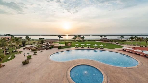 A resort pool area with a large central pool and a smaller circular pool, surrounded by lounge chairs and umbrellas, with a view of the ocean and sunset. - Radisson Blu Resort, Jizan (Jizan)