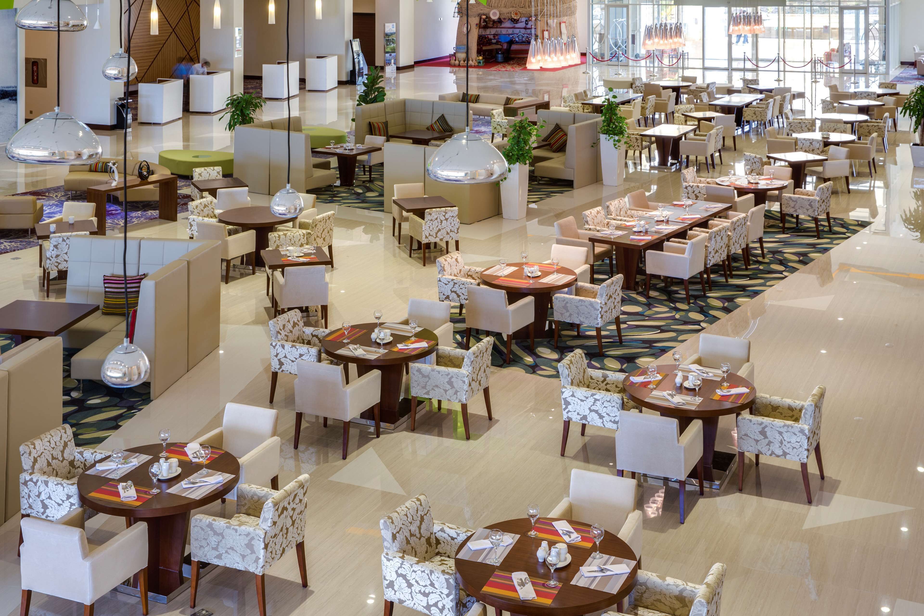 Un amplio vestíbulo de hotel con varias zonas de estar, incluyendo mesas redondas y sofás, y una zona central con bar.