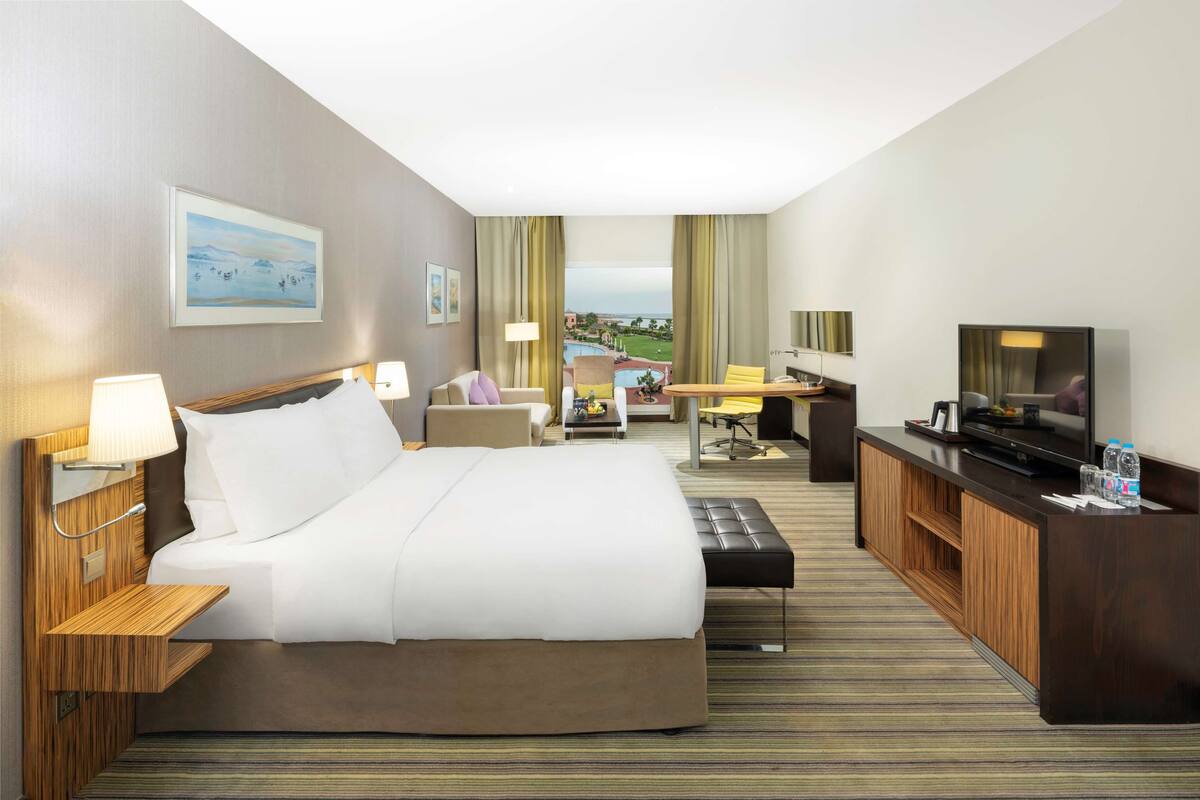 A hotel room with a large bed, a desk with a TV, a seating area with a view of the pool, and a balcony with curtains.