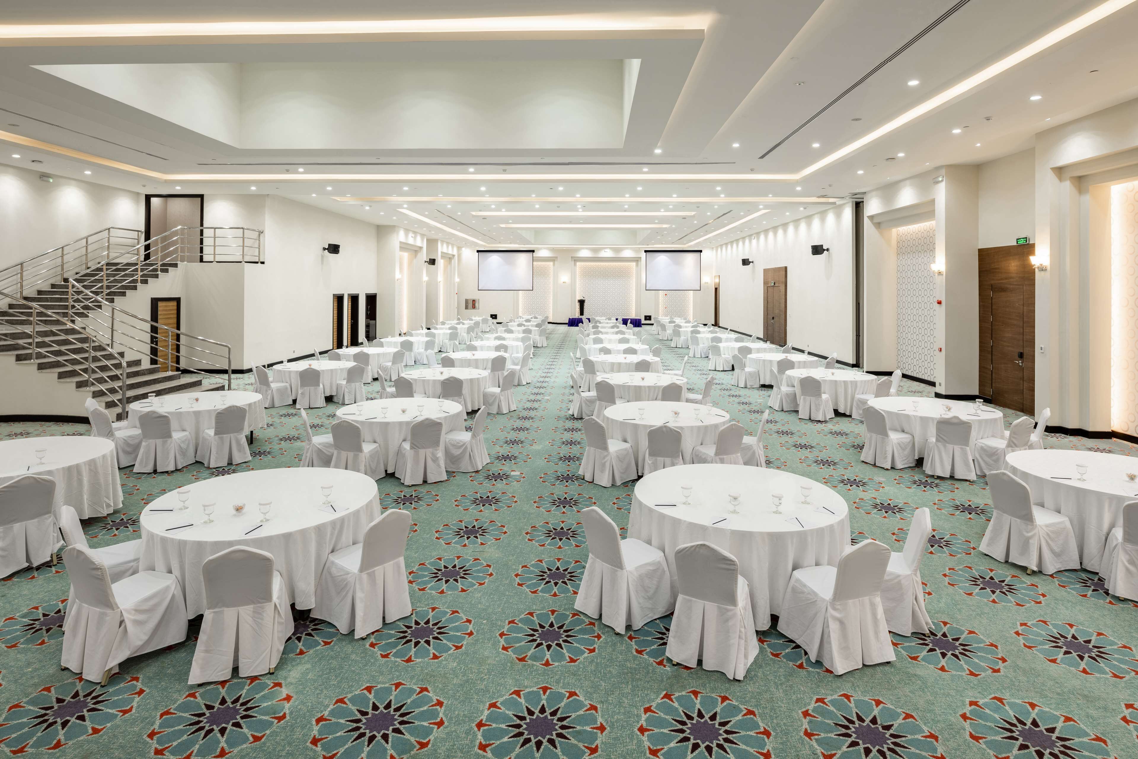 Un amplio salón de conferencias con varias mesas redondas, sillas blancas y suelo alfombrado.