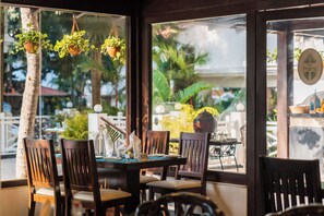 An indoor dining area with a table set for four, wooden chairs, hanging plants, and a view of palm trees and a building outside. - Radisson Goa Candolim (Candolim)