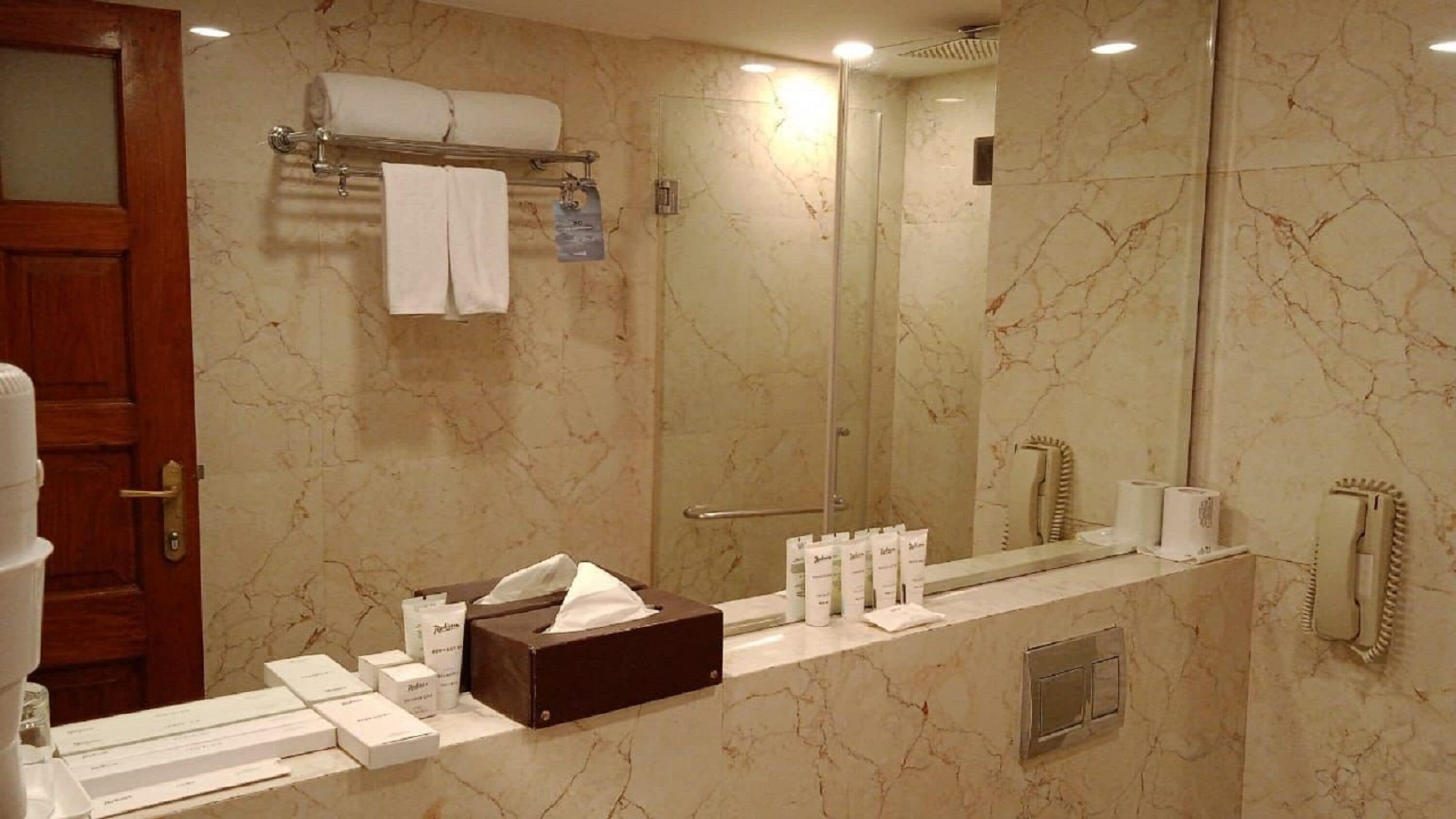 Een badkamer met een marmeren wand, een glazen douchecabine, een handdoekrek, een toilet en een wastafel met toiletartikelen.