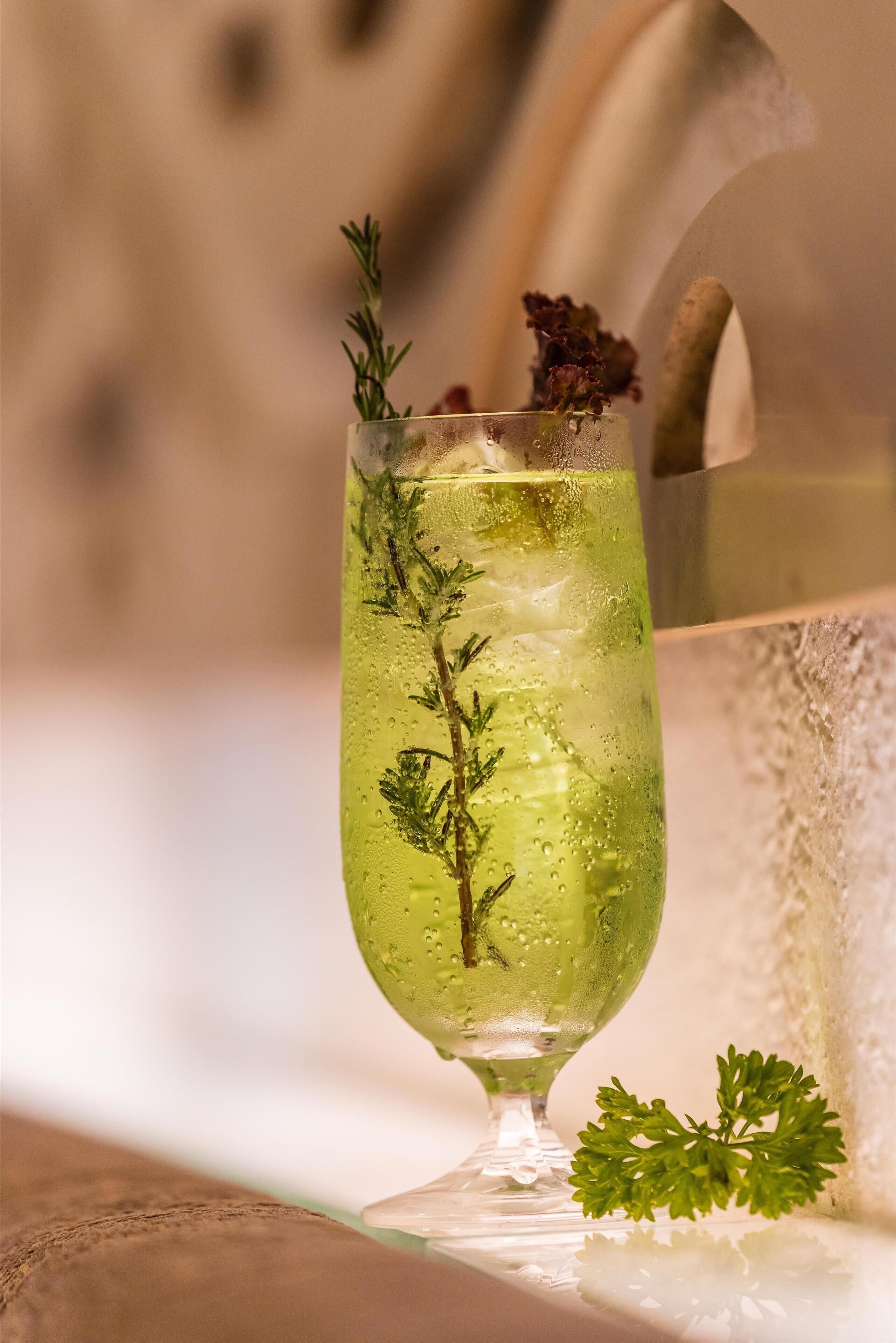 A glass of green beverage with herbs, ice, and a garnish, placed on a reflective surface with a blurred background.