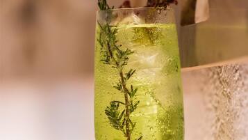 A glass of green beverage with herbs, ice, and a garnish, placed on a reflective surface with a blurred background.
