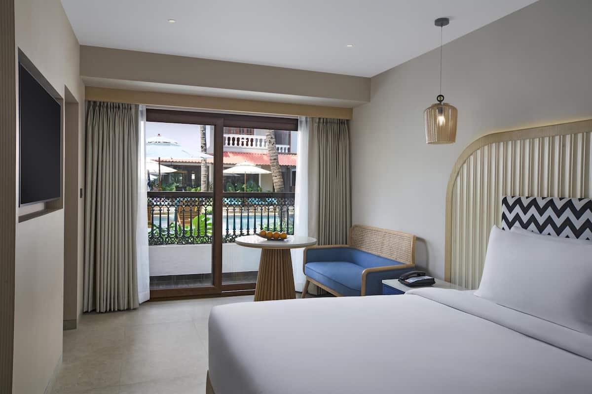 Habitación de hotel con cama, una mesita con fruta, una silla azul, balcón con vistas a edificios y una piscina, y una lámpara colgante.