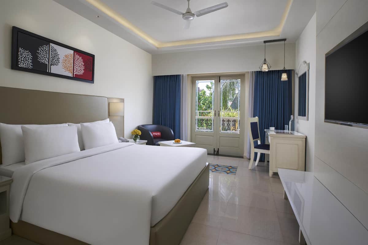 Habitación de hotel con una cama grande, un escritorio, una silla, un televisor, un ventilador de techo y un balcón con cortinas.