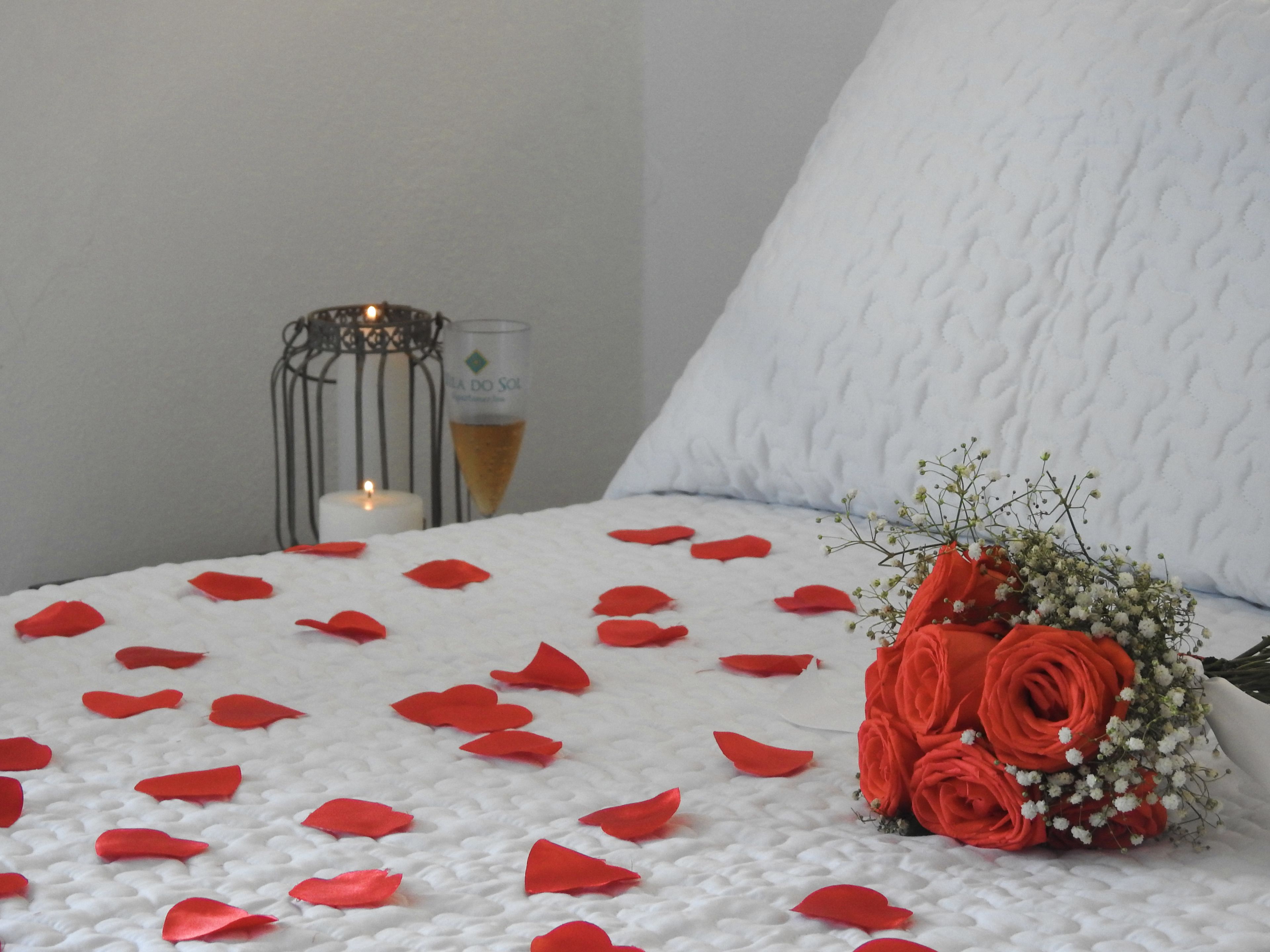 A bed with white bedding, red rose petals, a candle, and a glass of champagne.