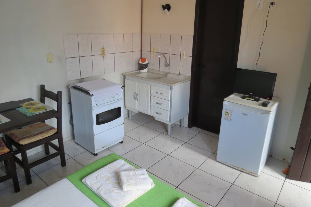A compact kitchen with a refrigerator, a sink, and a stove. There is a chair and a table with a mat.