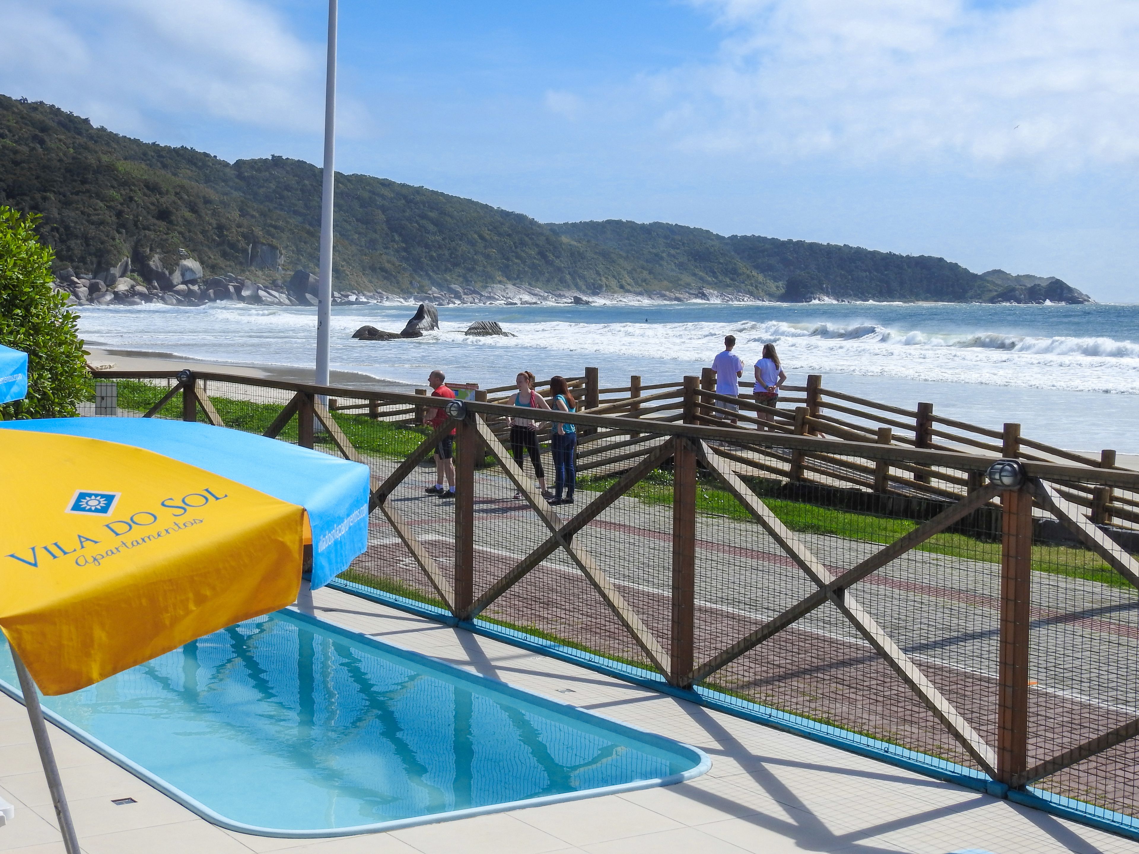 Terraza de madera con vistas a una playa con olas, piscina y un cartel de Apartamentos Vila do Sol.
