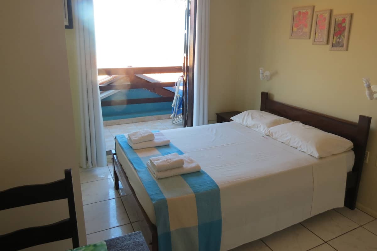 A hotel room with a bed, a small table, a chair, a window with curtains, and a view of a pool area.