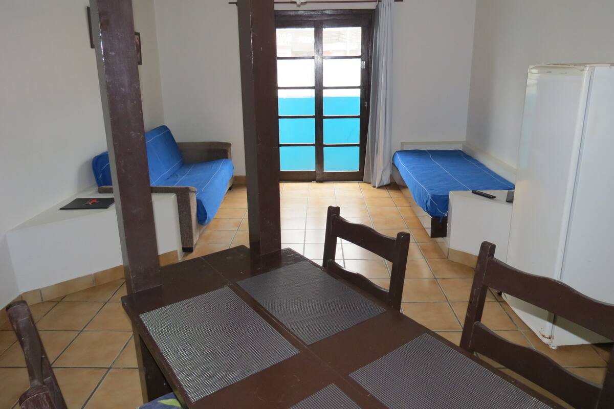 A room with a dining table, two blue beds, a ceiling fan, and a view of the sea through a large window.