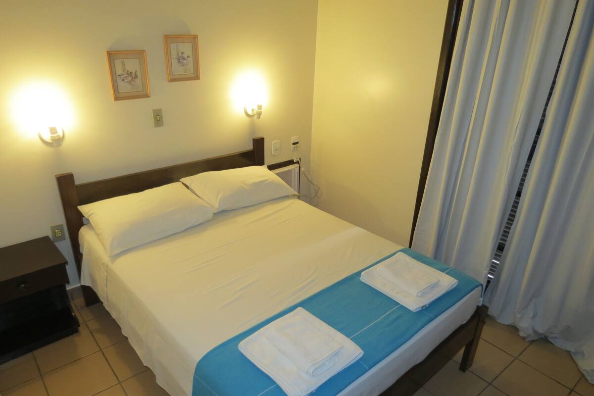 A hotel room with a bed, two bedside lamps, a nightstand, a window with curtains, and a wall with two framed pictures.
