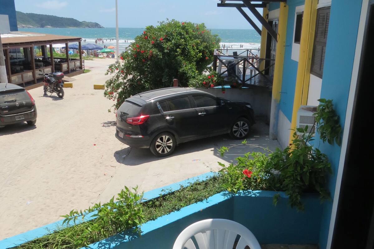 A balcony with a view of the beach, a parked car, and a restaurant with outdoor seating.