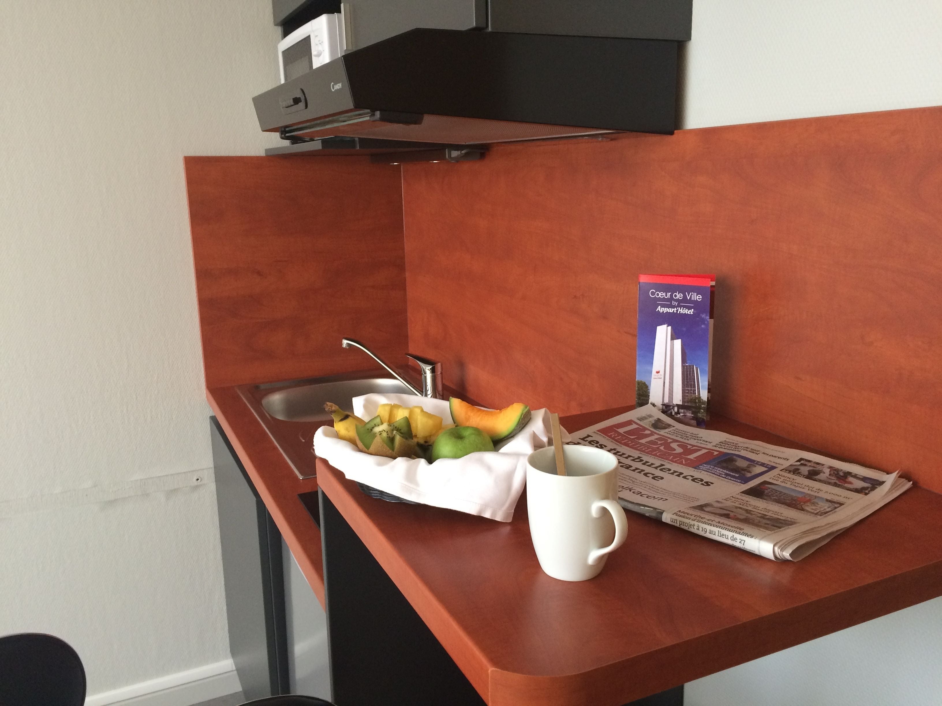 A small kitchen area with a sink, a white mug, a fruit bowl, and a book titled "Cœur de Ville" on a wooden counter.