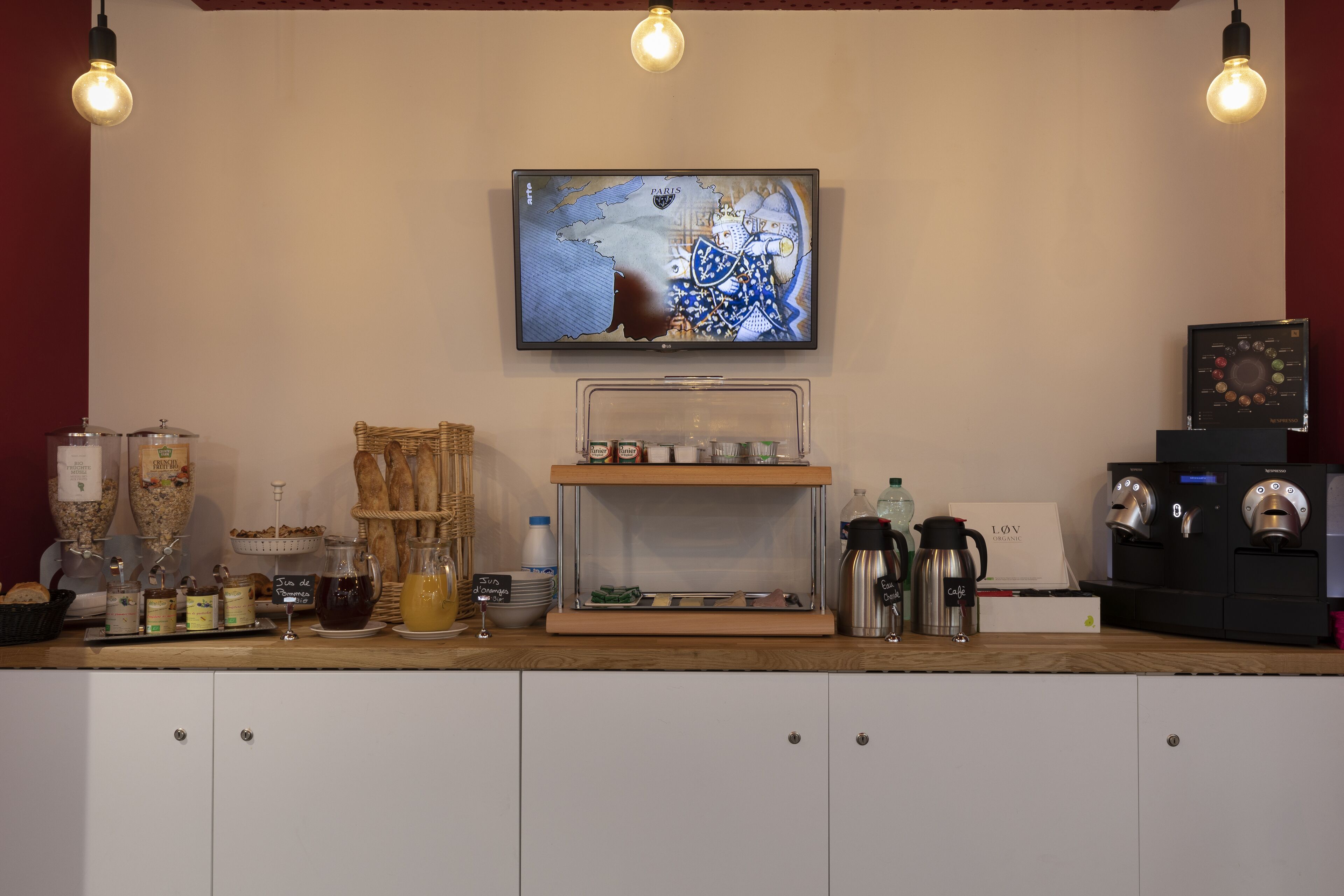 A café counter with a coffee machine, a television displaying a map, and various food items.