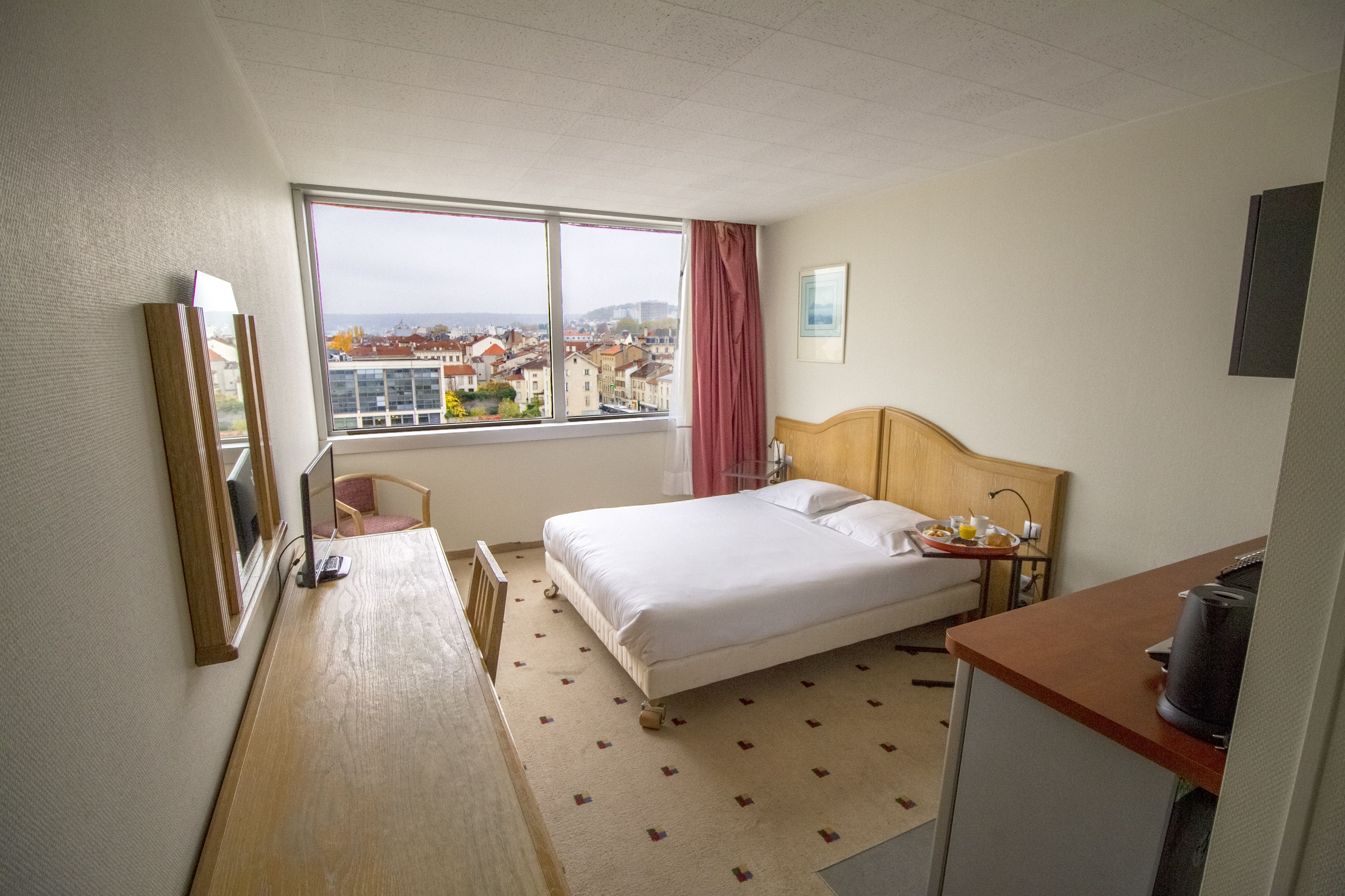 A hotel room with a bed, a desk, a chair, a window with a view of buildings, and a painting on the wall.
