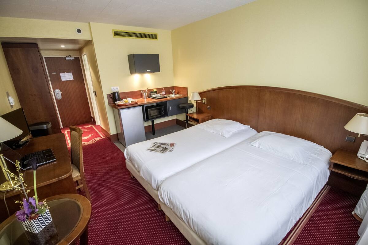 Habitación de hotel con una cama grande, un escritorio con computadora, una kitchenette pequeña y un televisor en la pared.