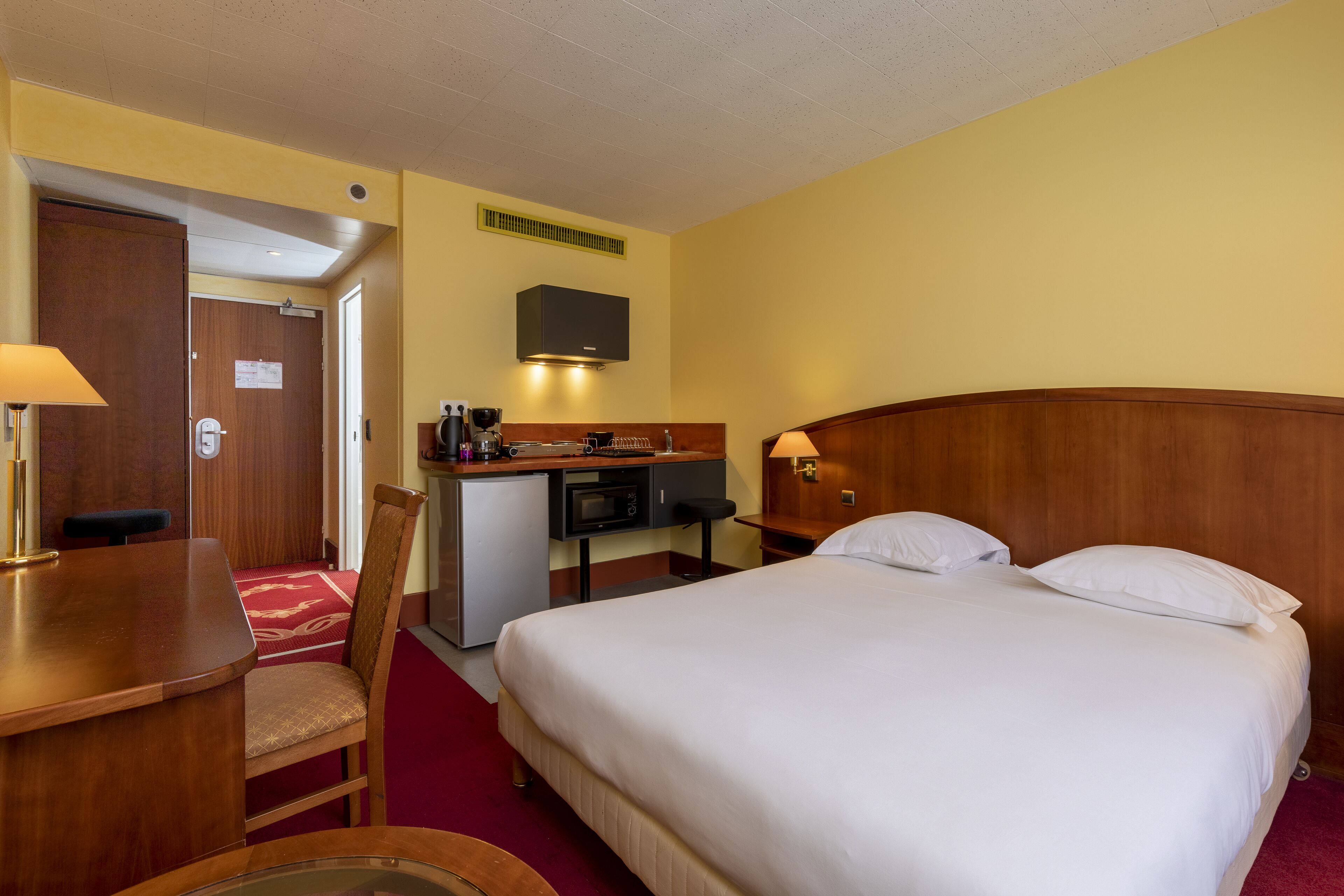 A hotel room with a bed, a desk, a chair, a small kitchenette, and a television.