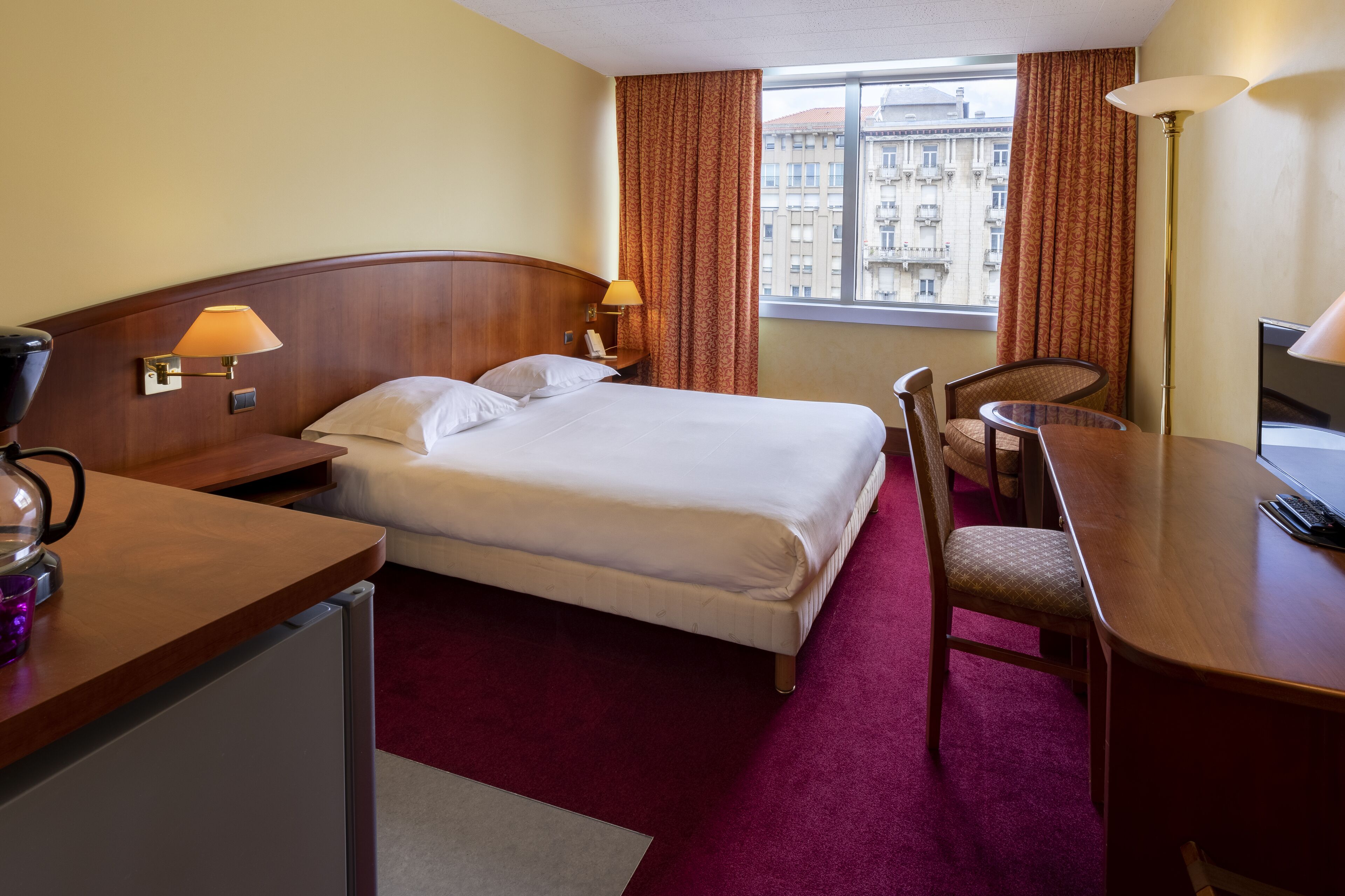 A hotel room with a bed, desk, chair, and a view of the city through the window.