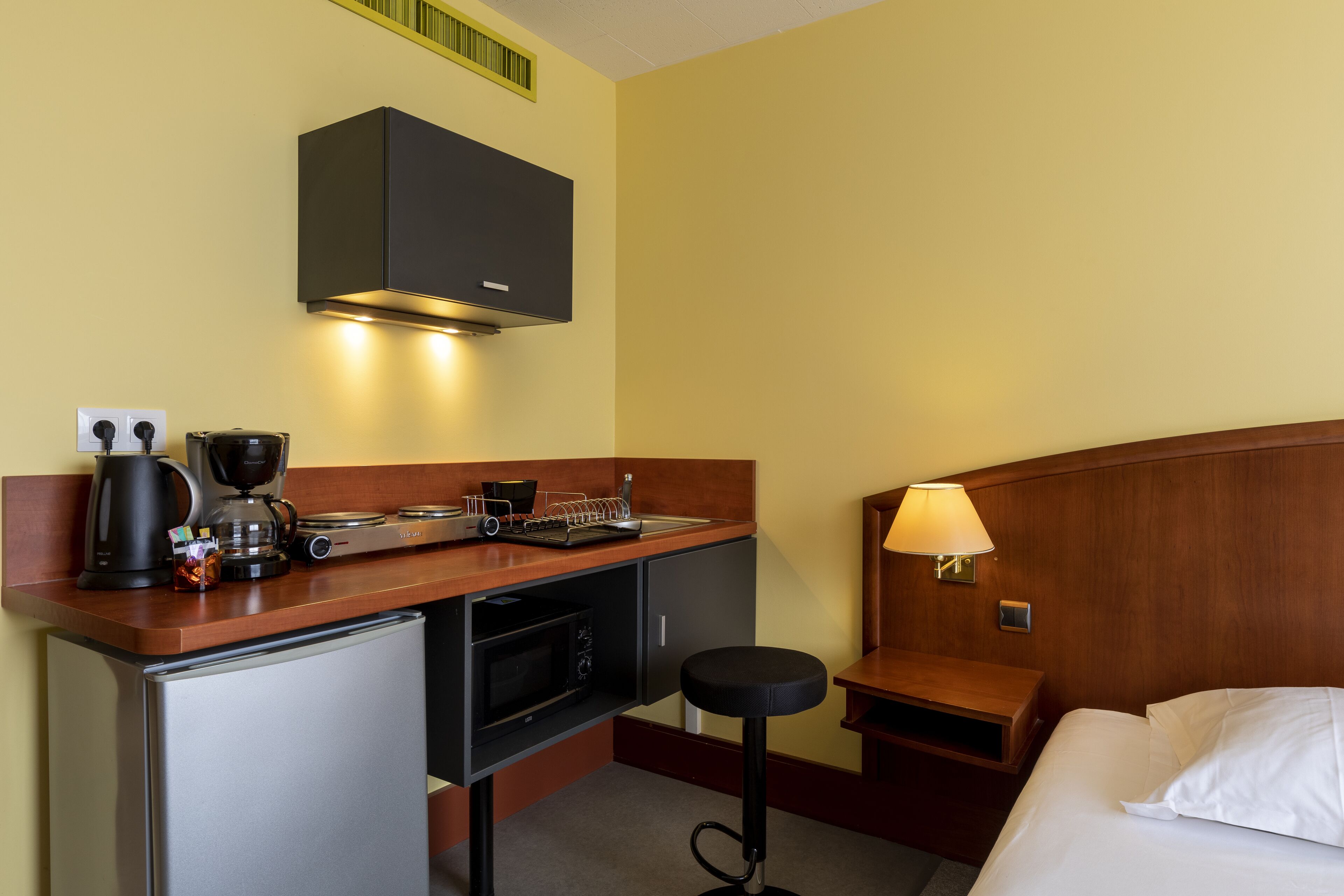 A compact hotel room with a kitchenette, a bed, a nightstand, and a small stool.